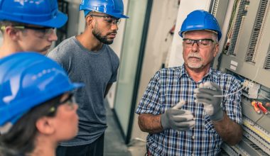Photo of a group of people in an industrial training setting wearing blue safety helmets. One man in a checkered shirt and gloves is demonstrating electrical work near a control panel while others observe attentively.