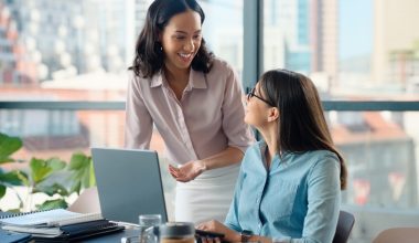 Two women collaborate at a modern office desk, with one standing and gesturing toward an open laptop while the other sits, listens, and learns. The workspace includes notebooks, a coffee cup, and a smartphone, and large windows behind them reveal a city skyline filled with natural light.