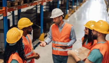 Photo of a group of workers in a warehouse wearing orange safety vests and yellow or white hard hats. A man in a white hard hat and orange vest stands at the center holding a tablet and speaking to the team. The others, including women and men, stand around him listening; one person on the right holds a cardboard box. Rows of shelves and inventory line the background.
