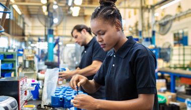 Photo of two assembly line workers in a factory setting; a foreground worker appearing female with hair in a braided bun wears a black short‑sleeve polo shirt and uses a hand tool to assemble blue mechanical components on a workbench, while a background worker appearing male in a black shirt performs a similar task along the same production line.