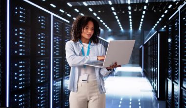 Photo: adult woman with curly dark hair wearing a light blue shirt, ID lanyard, and beige pants, using a laptop while standing in a data center aisle lined with illuminated server racks.